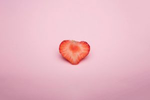 A heart-shaped strawberry slice on a soft pink background, highlighting its vivid red color.