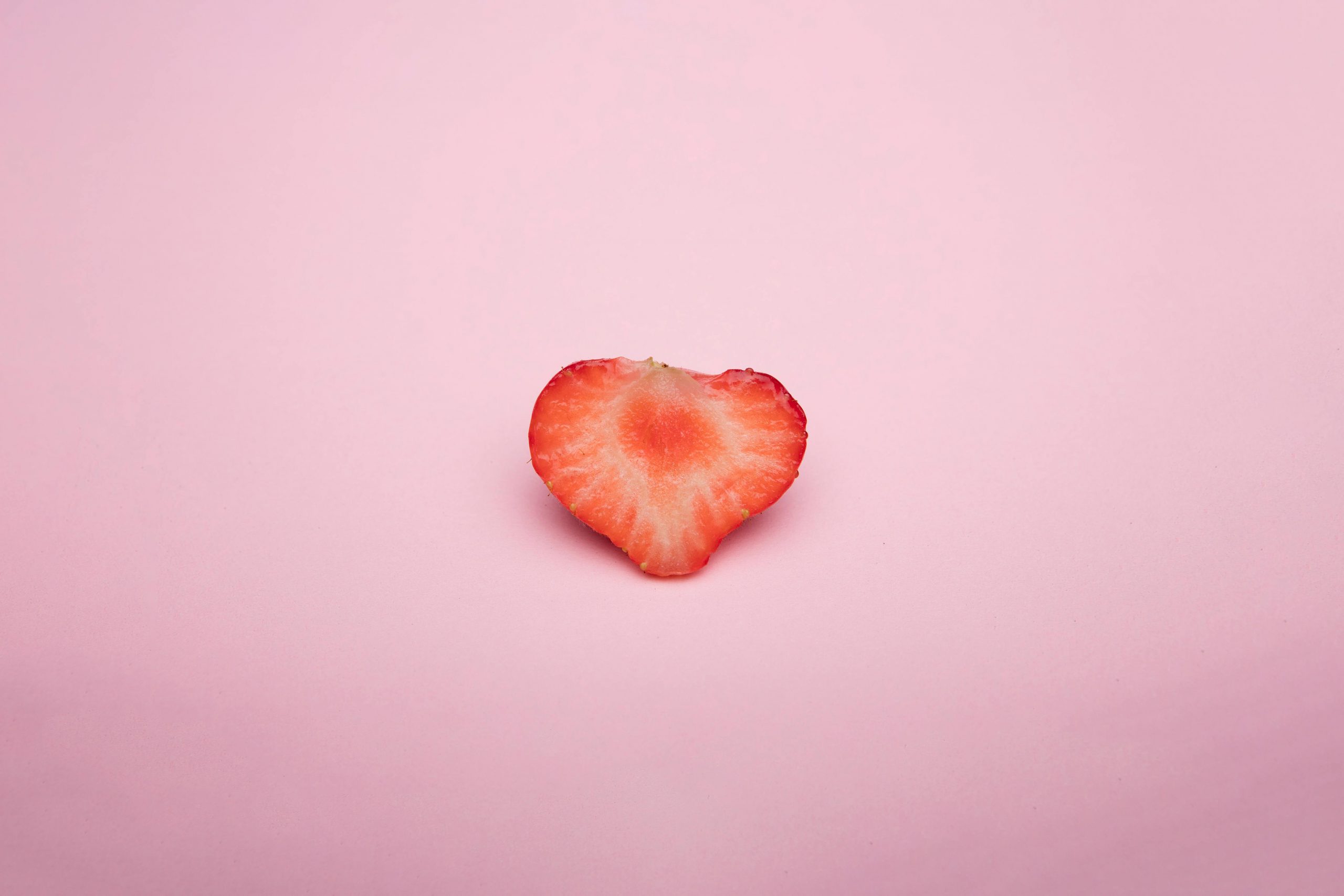 A heart-shaped strawberry slice on a soft pink background, highlighting its vivid red color.